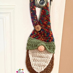 May include: A crocheted gnome door hanger with a multi-colored hat, green brim, and a brown beard. The gnome is hanging on a white door.