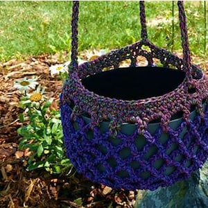 May include: A hanging crochet plant basket in shades of purple, green, and brown. The basket has a woven design and a sturdy handle for easy hanging. The basket is empty and ready for a plant.