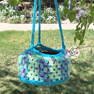 May include: A crocheted hanging planter in shades of blue, green, and purple. The planter has a round shape and is made with a textured stitch. The planter is hanging from a tree branch.
