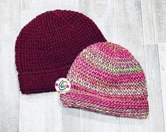 May include: Two knitted beanies. One is a solid burgundy color, and the other is a mix of pink, green, and cream. Both beanies have a ribbed brim and a textured knit pattern. The beanies are likely handmade and suitable for cold weather.
