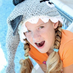 Può includere: Una ragazza con un cappello di squalo in crochet grigio con denti bianchi e una benda nera sull'occhio. Indossa una camicia arancione brillante e ha i capelli biondi in due trecce.