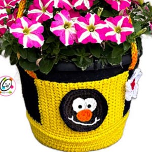 May include: A black flower pot with a yellow and black crocheted cover featuring a cartoon face. The pot is filled with vibrant pink and white petunia flowers. The crocheted cover has a round black face with white eyes and an orange nose.
