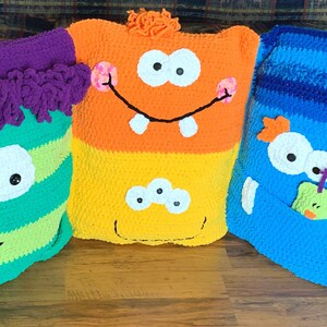 May include: Three crocheted monster pillows. The first pillow is green and yellow striped with a purple hair tuft, the second is orange and yellow with a white eye and teeth, and the third is blue with a green monster applique.