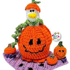 May include: A crocheted pumpkin with a smiling face, wearing a green scarf and a white crocheted hat with a yellow beak. There are three smaller pumpkins on a purple crocheted mat with black spiders.