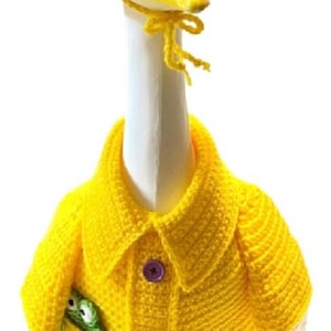 Op de afbeelding: Een grillig, handgemaakt ganzenbeeldje met een felgele gehaakte hoed en jas. De jas heeft paarse knopen en een zak met een groene kikker. De gans heeft een witte nek en een gele snavel.