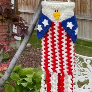 May include: A crocheted American flag-themed eagle-shaped bag with red, white, and blue stripes and stars. The bag is hanging from a tree branch.