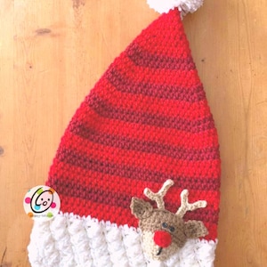 Puede incluir: Sombrero de Papá Noel de punto rojo y blanco con un pompón blanco en la parte superior y una cara de reno de punto marrón en la parte delantera. El sombrero tiene un borde de punto blanco.
