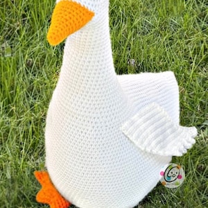 May include: White crocheted goose with an orange beak and feet. The goose is sitting on a bed of green grass.