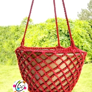 May include: A red crocheted hanging planter with a diamond pattern. The planter is suspended by three red cords, with a light brown pot visible inside. The background features green foliage and a bright sky.