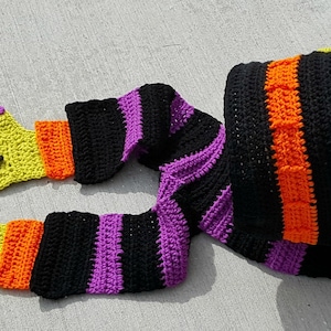 May include: A whimsical, crocheted Halloween costume piece. It features a black hood with orange and purple stripes, and star accents. The arms are striped black and purple, with yellow hands, purple fingernails, and black spider accents.