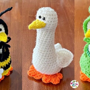 May include: Three crocheted goose toys wearing different costumes. One goose is wearing a black and yellow bee costume, another is wearing a white dress, and the third is wearing a green frog costume.