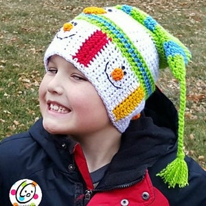 Pode incluir: Um chapéu de inverno de crochê colorido com um design de boneco de neve. O chapéu é principalmente branco com detalhes em vermelho, amarelo, verde e azul. Possui uma longa borla verde e é usado por uma pessoa.