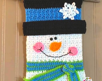 Peut inclure: Une décoration murale en crochet représentant un bonhomme de neige avec un chapeau noir, une écharpe bleu et verte et un flocon de neige blanc. Le bonhomme de neige a un corps blanc, des yeux noirs, un nez orange et des joues roses.
