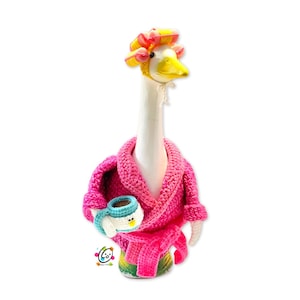 May include: A whimsical goose figurine with a white neck and yellow beak, wearing a pink crocheted robe and hair curlers. The goose holds a small, blue and white teacup. The figurine is a fun, decorative item.