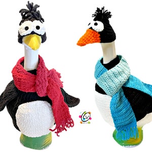 May include: Two crocheted goose figurines wearing winter hats and scarves. One goose is wearing a black penguin hat with a red scarf, and the other is wearing a black hat with an orange beak and a blue scarf.