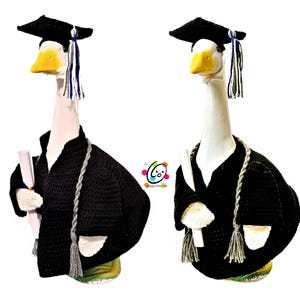 May include: Two goose ornaments, each wearing a black graduation gown and mortarboard cap with a blue tassel. One goose holds a rolled diploma. The geese have yellow beaks and are wearing grey braided cords.