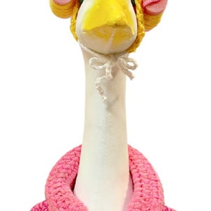 May include: A white goose figurine with a yellow beak and black eyes, wearing a yellow crocheted cap with pink foam curlers. The goose is dressed in a pink crocheted robe, tied with a white bow.