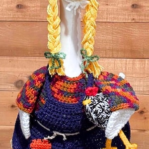 May include: A whimsical goose figurine dressed in a handmade crochet outfit. The goose wears a blue hat with a yellow flower, a blue dress, and yellow braids. It holds a small crochet chicken purse. The outfit features orange, red, and green accents.