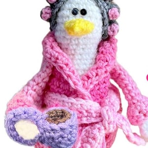 May include: A handmade crochet penguin wearing a pink robe and curlers. The penguin is holding a purple mug and has orange feet. The penguin's robe is a gradient of pinks, and the curlers are gray with pink accents.