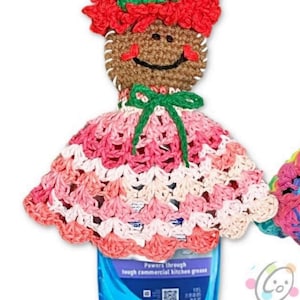 Puede incluir: Una funda de muñeca de ganchillo hecha a mano con forma de hombre de jengibre, con pelo de hilo rojo, un lazo verde y un vestido rosa y blanco. La funda de la muñeca se coloca sobre una botella azul.