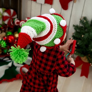 May include: A child wearing a red, white, and green striped elf hat with a green pom-pom. The hat has white pom-poms around the brim.