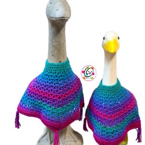 May include: Two goose statues wearing colorful, handmade crochet ponchos. The ponchos feature a vibrant gradient of blue, purple, and pink, with matching fringe tassels. The statues are set against a white background.