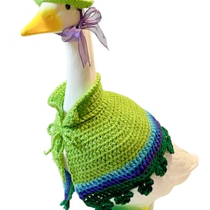 May include: A white goose figurine wearing a green crocheted hat and a green crocheted cape with purple and blue accents. The cape has a clover design at the bottom.