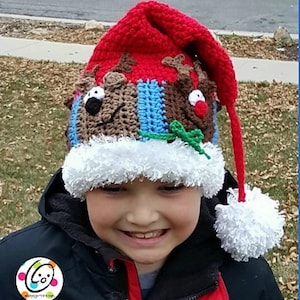 Pode incluir: Uma criança usando um chapéu de Papai Noel de crochê vermelho e branco com um design de rena. O chapéu tem um pompom branco na ponta da longa borla vermelha. O chapéu é feito com uma variedade de cores, incluindo vermelho, branco, azul, verde e marrom.