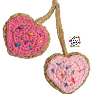 May include: Two pink heart-shaped crocheted cookies with brown edges and colorful sprinkles. The cookies are connected by a brown crocheted string.