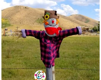 May include: A scarecrow with a red and black plaid shirt, a brown hat with sunflowers, and a red scarf stands in a field with a wooden post and a barbed wire fence.