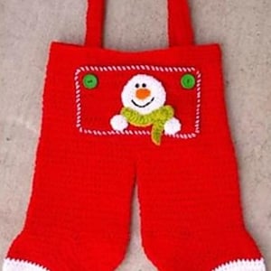 May include: Red crocheted Christmas stocking shaped like overalls, featuring a snowman applique with a green scarf and two green buttons. The stocking has white cuffs and suspenders.