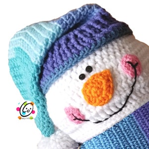 Könnte beinhalten: Ein gehäkelter Schneemann mit einem blau-weiß gestreiften Hut, einer orangenen Nase und rosafarbenen Wangen. Der Schneemann lächelt.
