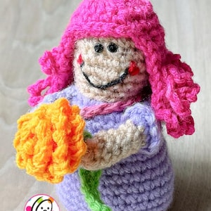Op de afbeelding: Een gehaakte pop met roze haar en een paarse jurk die een gele bloem vasthoudt.