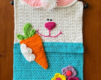 Pode incluir: Um coelho de crochê branco com orelhas cor-de-rosa e um nariz de pompom branco segurando uma cenoura. O coelho está preso a um bolso de crochê azul com três flores de crochê amarelas, cor-de-rosa e roxas.