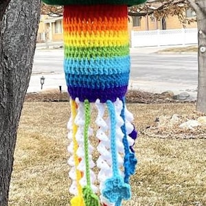 May include: A whimsical, handmade crochet wind chime. The top is a green and black leprechaun hat with a gold buckle. The body is a rainbow of colors, and the bottom has colorful, dangling accents. A unique, decorative item.