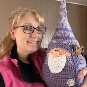 May include: A woman is holding a purple and white crocheted gnome with a brown crocheted mouse on its back. The gnome has a white beard and a pink nose.