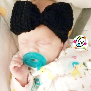 May include: A baby wearing a black crocheted bow headband and a white onesie with a colorful animal print. The baby is holding a blue pacifier.