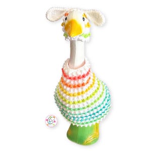 Könnte beinhalten: Eine weiße gehäkelte Gans mit einem regenbogenfarbenen gestreiften Pullover und einer weißen gehäkelten Mütze mit Ohren.