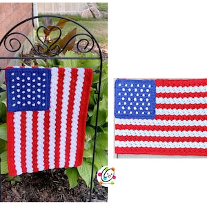 May include: A crocheted American flag with red, white, and blue yarn. The flag is hanging on a black metal stand in front of a brick wall and green plants.