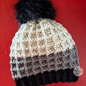 May include: A handmade crochet beanie in a waffle stitch pattern. The hat features a black pom-pom and is striped with shades of gray and black. The ribbed brim is black. A small, round sticker is on the side of the hat.