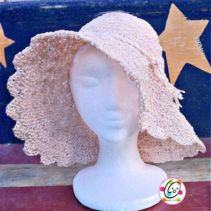 Puede incluir: Un sombrero de sol de crochet color melocotón claro con un ala ancha y festoneada. El sombrero se exhibe en una cabeza de maniquí blanca. La textura del sombrero es visible y el ala tiene un borde decorativo. El sombrero está diseñado para proteger del sol.
