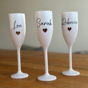 Könnte beinhalten: Drei weiße Champagnerflöten mit goldenen Herzen und schwarzen Kalligraphie-Namen: Lea, Sarah und Rebecca.