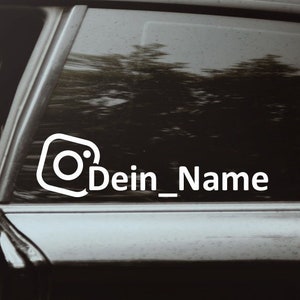 Könnte beinhalten: Weißer Aufkleber mit Instagram-Logo und dem Text "Dein_Name" auf einem Autofenster. Das Design ist schlicht und modern. Der Hintergrund zeigt eine Person im Auto. Ideal zur Fahrzeugindividualisierung.