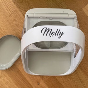 May include: A white and gray automatic pet feeder with a curved handle and a removable tray. The name "Molly" is printed in black script on the front. The feeder is designed to dispense food for pets.