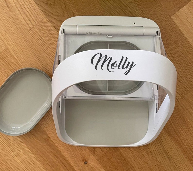 May include: A white and gray automatic pet feeder with a curved handle and a removable tray. The name "Molly" is printed in black script on the front. The feeder is designed to dispense food for pets.