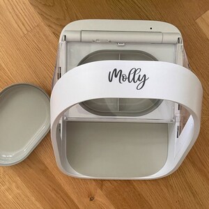 May include: A white and gray automatic pet feeder with a removable tray. The feeder has a curved handle and the name "Molly" in script. The tray is oval-shaped and sits next to the feeder on a wooden surface.