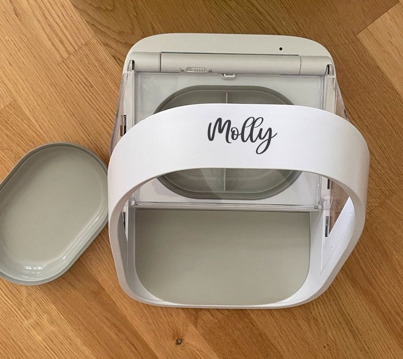 May include: A white and gray automatic pet feeder with a removable tray. The feeder has a curved handle and the name "Molly" in script. The tray is oval-shaped and sits next to the feeder on a wooden surface.