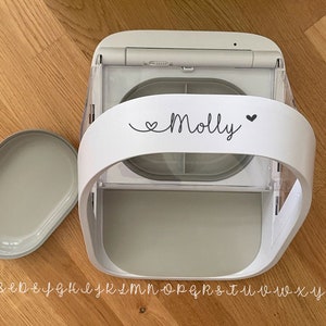 May include: A white and gray automatic pet feeder with a removable lid. The feeder has a curved handle with the name "Molly" and a heart symbol printed on it. The feeder is on a wooden surface.