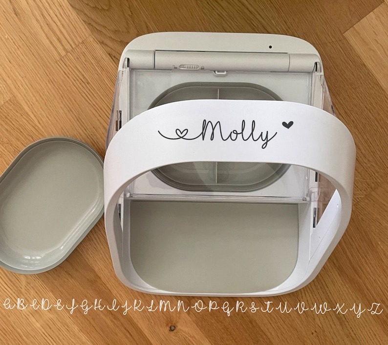 May include: A white and gray automatic pet feeder with a removable lid. The feeder has a curved handle with the name "Molly" and a heart symbol printed on it. The feeder is on a wooden surface.
