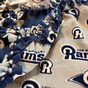 May include: A blue and gray fleece blanket featuring the Los Angeles Rams logo and team name. The blanket has a knotted edge in blue and white, and is made of soft fleece material.